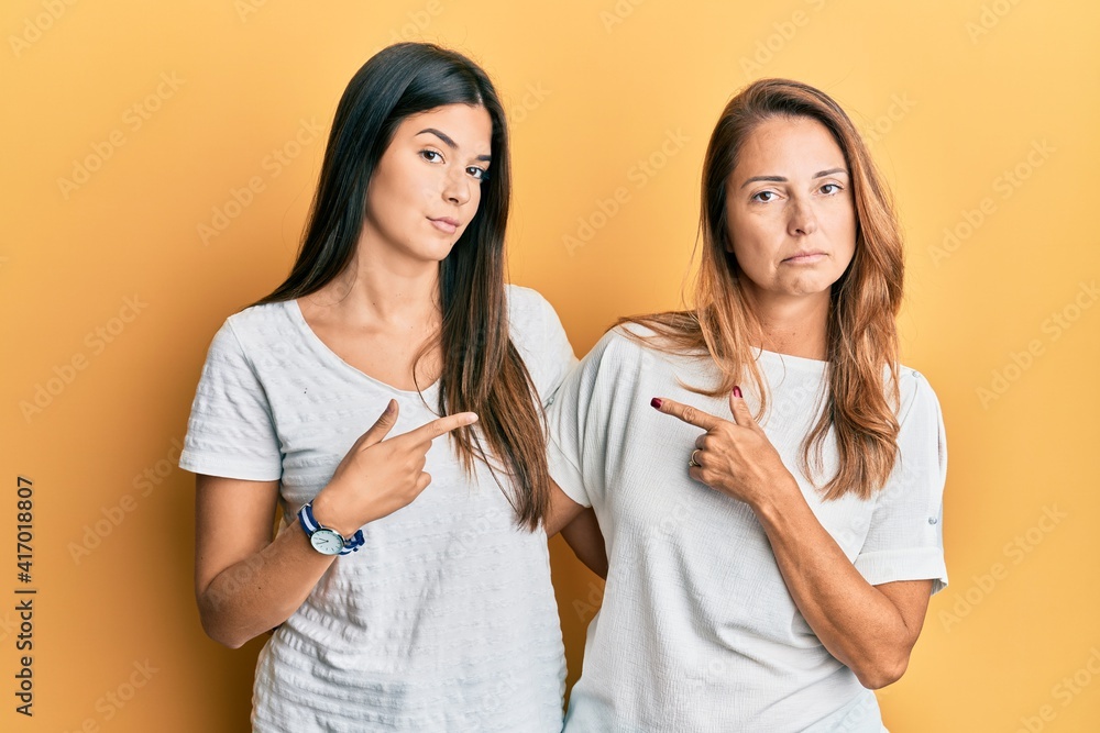 Hispanic family of mother and daughter wearing casual white tshirt pointing with hand finger to the side showing advertisement, serious and calm face Hispanic family of mother and daughter wearing casual white tshirt pointing with hand finger to the side showing advertisement, serious and calm face