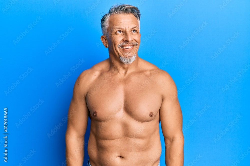 Fototapeta premium Middle age grey-haired man standing shirtless looking away to side with smile on face, natural expression. laughing confident.