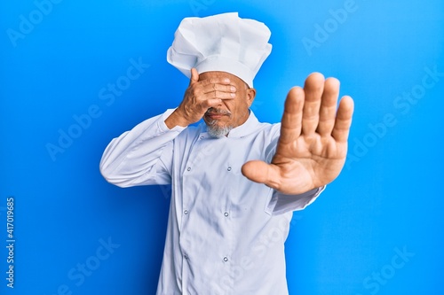 Middle age grey-haired man wearing professional cook uniform and hat covering eyes with hands and doing stop gesture with sad and fear expression. embarrassed and negative concept.