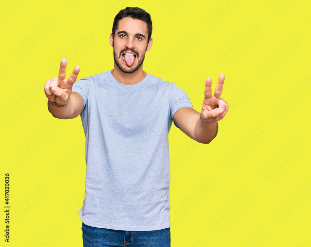 Young hispanic man wearing casual clothes smiling with tongue out showing fingers of both hands doing victory sign. number two.