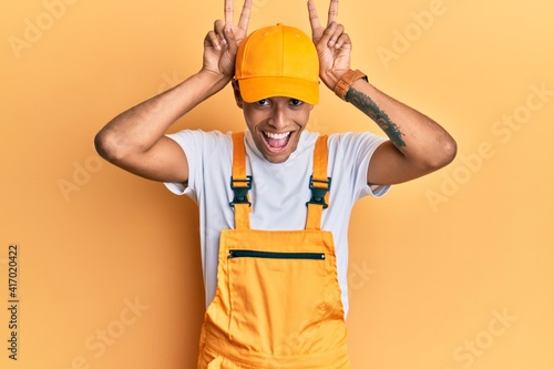 Young handsome african american man wearing handyman uniform over yellow background posing funny and crazy with fingers on head as bunny ears, smiling cheerful