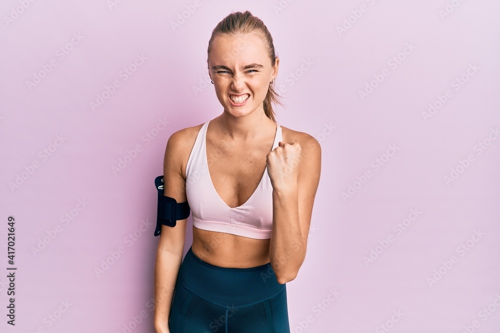 Beautiful blonde woman wearing sportswear and arm band angry and mad raising fist frustrated and furious while shouting with anger. rage and aggressive concept.