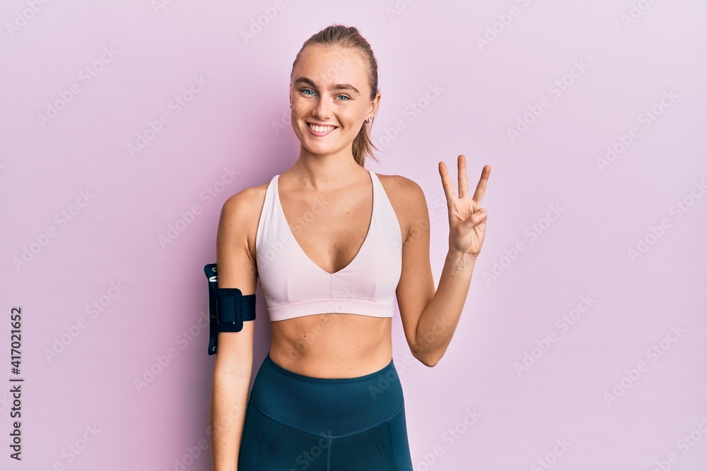 Beautiful blonde woman wearing sportswear and arm band showing and pointing up with fingers number three while smiling confident and happy.