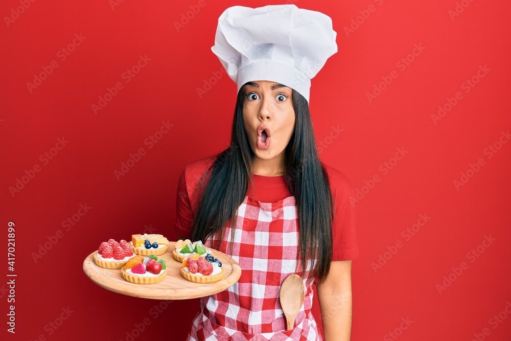 Young beautiful hispanic girl wearing baker apron holding cakes scared and amazed with open mouth for surprise, disbelief face Young beautiful hispanic girl wearing baker apron holding cakes scared and amazed with open mouth for surprise, disbelief face
