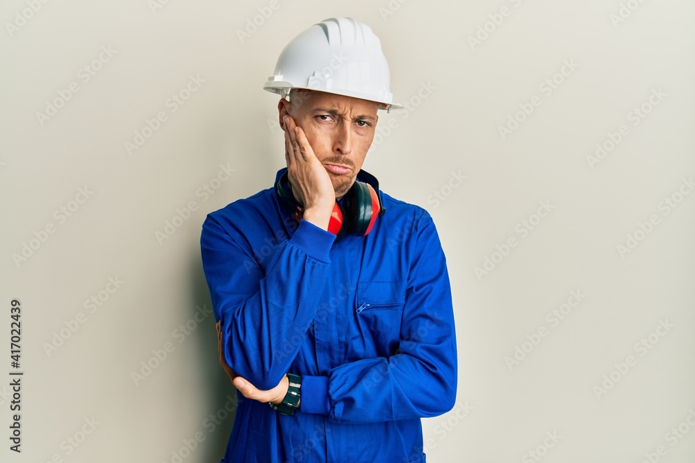 Bald man with beard wearing builder jumpsuit uniform and hardhat thinking looking tired and bored with depression problems with crossed arms.