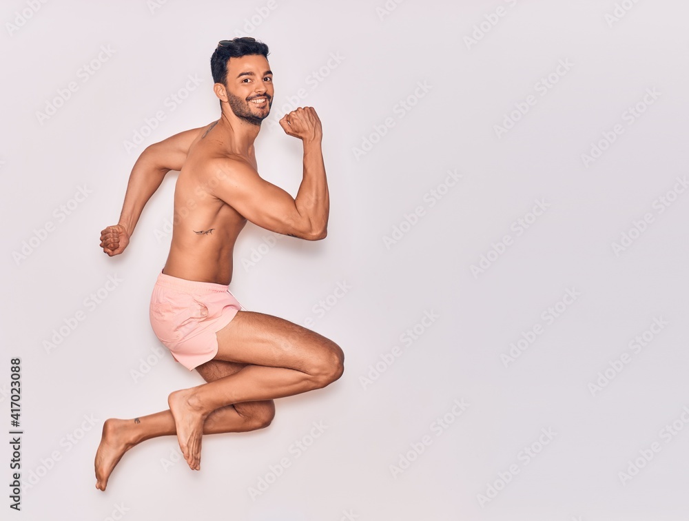 Young handsome hispanic man on vacation wearing swimwear shirtless smiling happy. Jumping with smile on face celebrating with fists up over isolated white background
