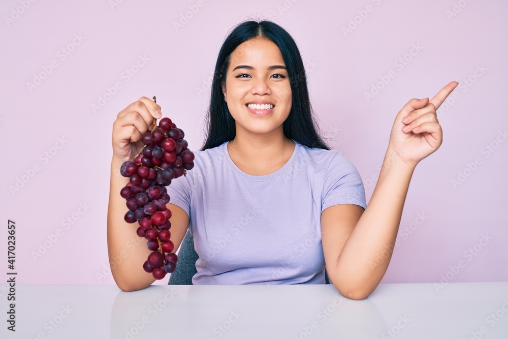 Young beautiful asian girl holding branch of fresh grapes smiling happy pointing with hand and finger to the side Young beautiful asian girl holding branch of fresh grapes smiling happy pointing with hand and finger to the side