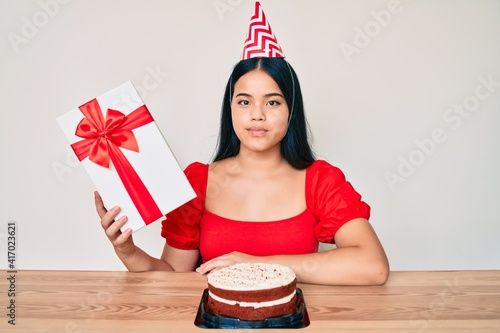 Fotografie Young asian girl wearing birthday hat holding present thinking attitude and sobe