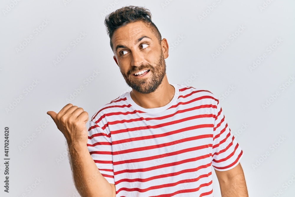 Handsome man with beard wearing casual clothes smiling with happy face looking and pointing to the side with thumb up. Handsome man with beard wearing casual clothes smiling with happy face looking and pointing to the side with thumb up.