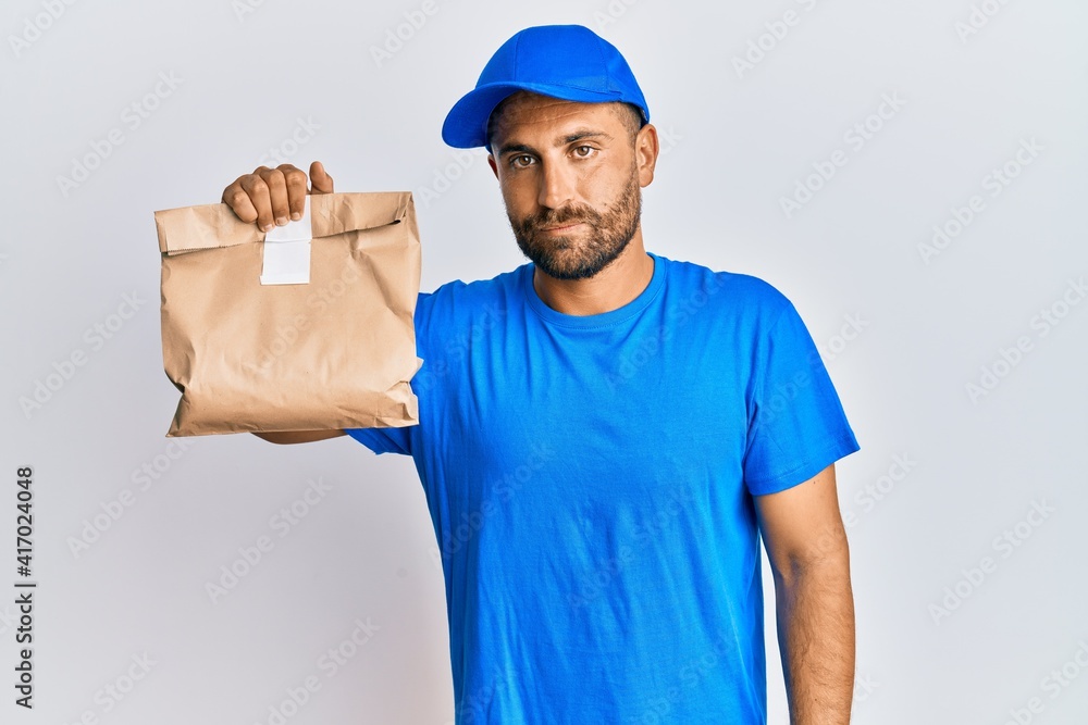 Handsome man with beard holding delivery package thinking attitude and sober expression looking self confident Handsome man with beard holding delivery package thinking attitude and sober expression looking self confident