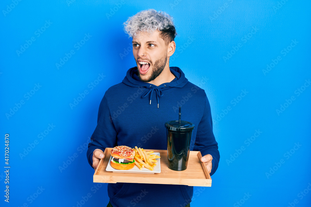 Young hispanic man with modern dyed hair eating a tasty classic burger with fries and soda angry and mad screaming frustrated and furious, shouting with anger. rage and aggressive concept. Young hispanic man with modern dyed hair eating a tasty classic burger with fries and soda angry and mad screaming frustrated and furious, shouting with anger. rage and aggressive concept.