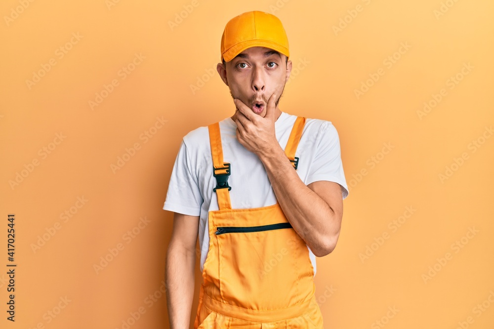 Hispanic young man wearing handyman uniform looking fascinated with disbelief, surprise and amazed expression with hands on chin Hispanic young man wearing handyman uniform looking fascinated with disbelief, surprise and amazed expression with hands on chin
