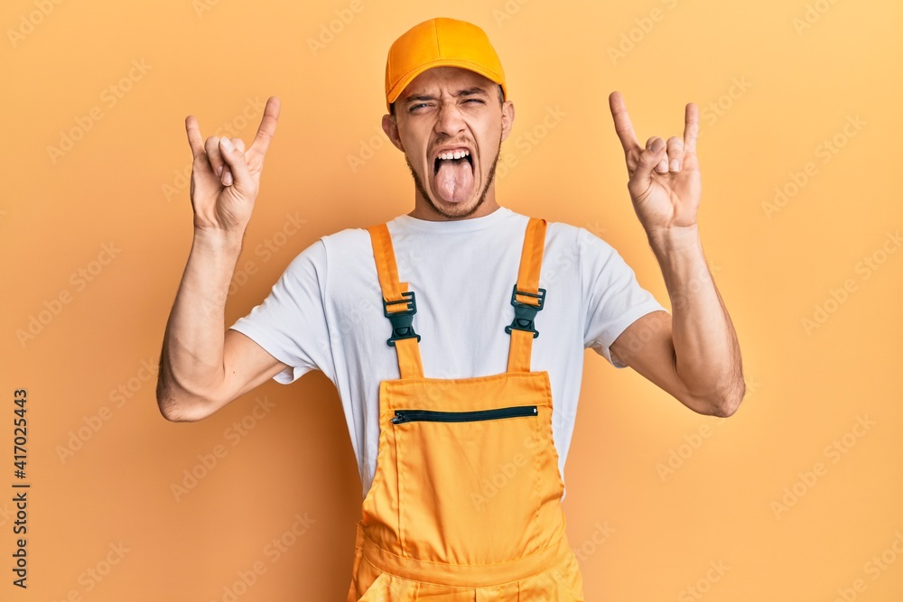 Hispanic young man wearing handyman uniform shouting with crazy expression doing rock symbol with hands up. music star. heavy music concept. Hispanic young man wearing handyman uniform shouting with crazy expression doing rock symbol with hands up. music star. heavy music concept.