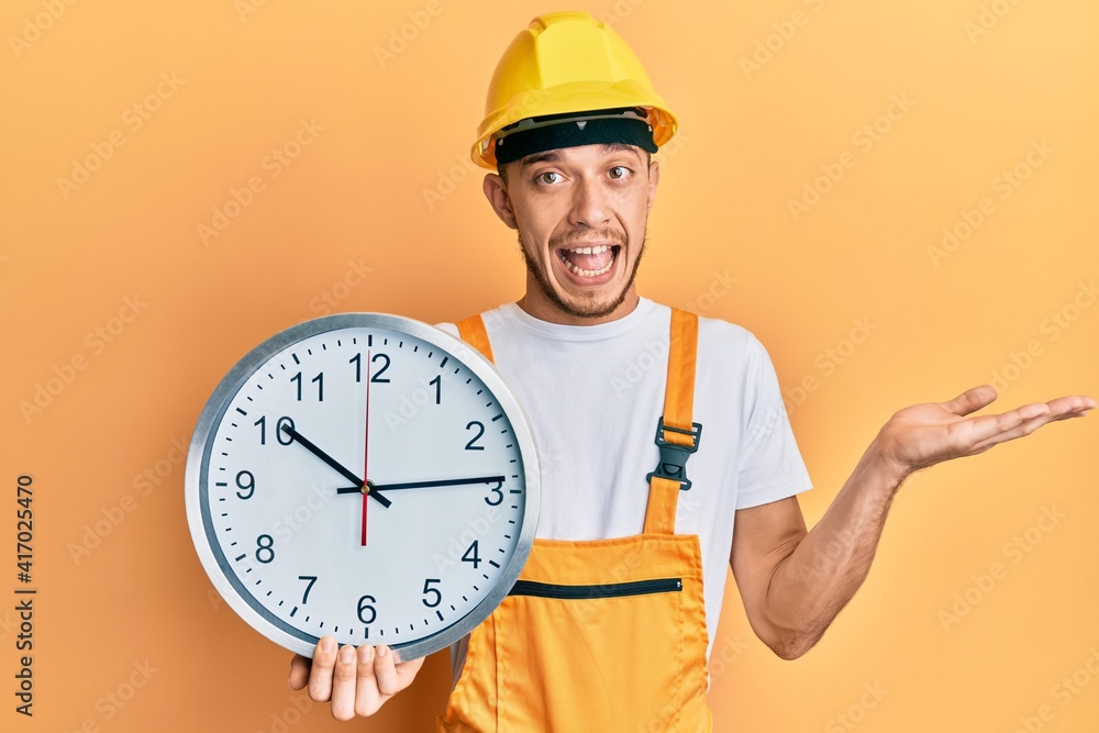 Hispanic young man wearing builder uniform and hardhat holding clock celebrating achievement with happy smile and winner expression with raised hand Hispanic young man wearing builder uniform and hardhat holding clock celebrating achievement with happy smile and winner expression with raised hand