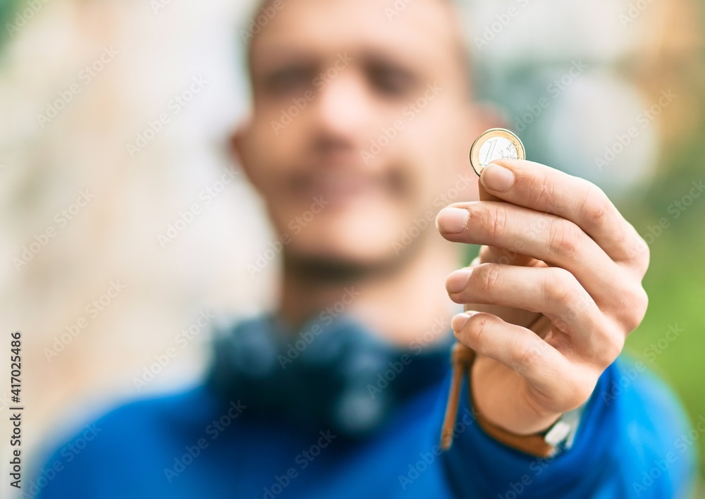 Young hispanic man using headphones holding 1 euro coin at the city