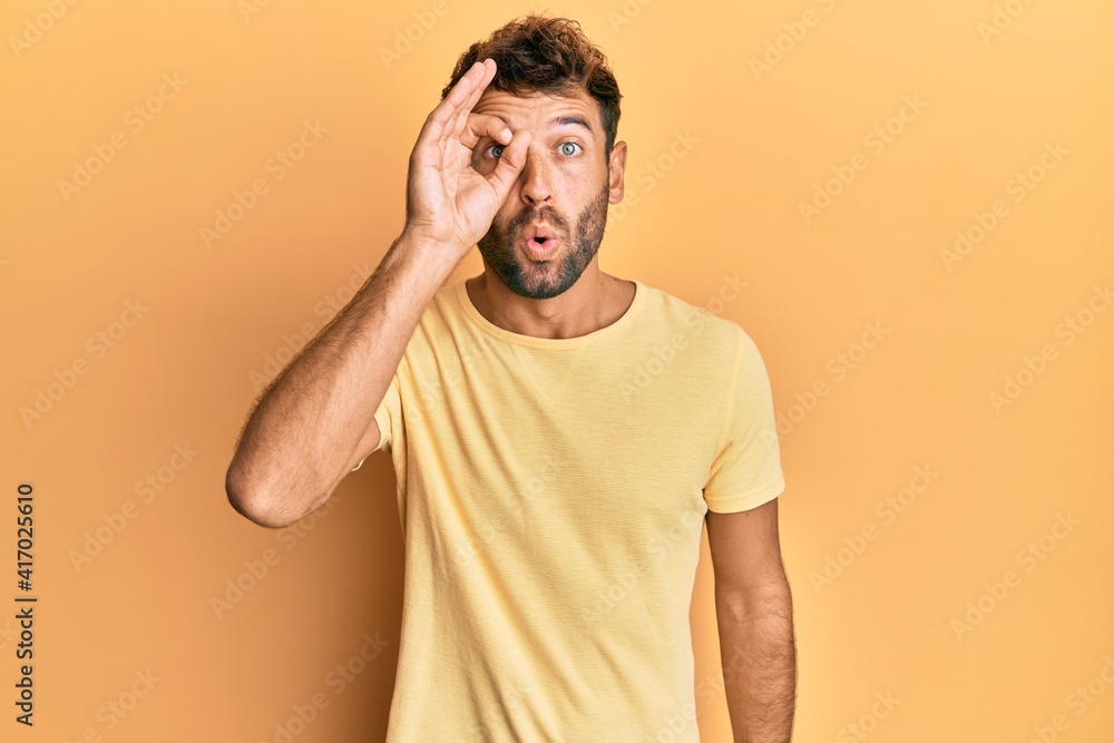 Handsome man with beard wearing casual yellow tshirt over yellow background doing ok gesture shocked with surprised face, eye looking through fingers. unbelieving expression. Handsome man with beard wearing casual yellow tshirt over yellow background doing ok gesture shocked with surprised face, eye looking through fingers. unbelieving expression.