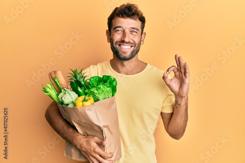 Handsome man with beard holding paper bag with bread and groceries doing ok sign with fingers, smiling friendly gesturing excellent symbol