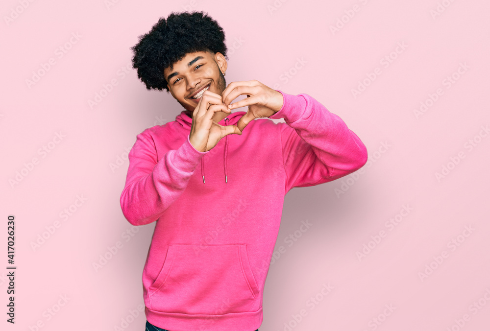 Young african american man with afro hair wearing casual pink sweatshirt smiling in love doing heart symbol shape with hands. romantic concept. Young african american man with afro hair wearing casual pink sweatshirt smiling in love doing heart symbol shape with hands. romantic concept.