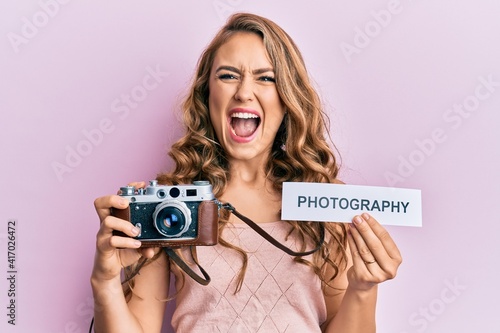 Young blonde girl holding vintage camera and paper with photography word paper smiling and laughing hard out loud because funny crazy joke.