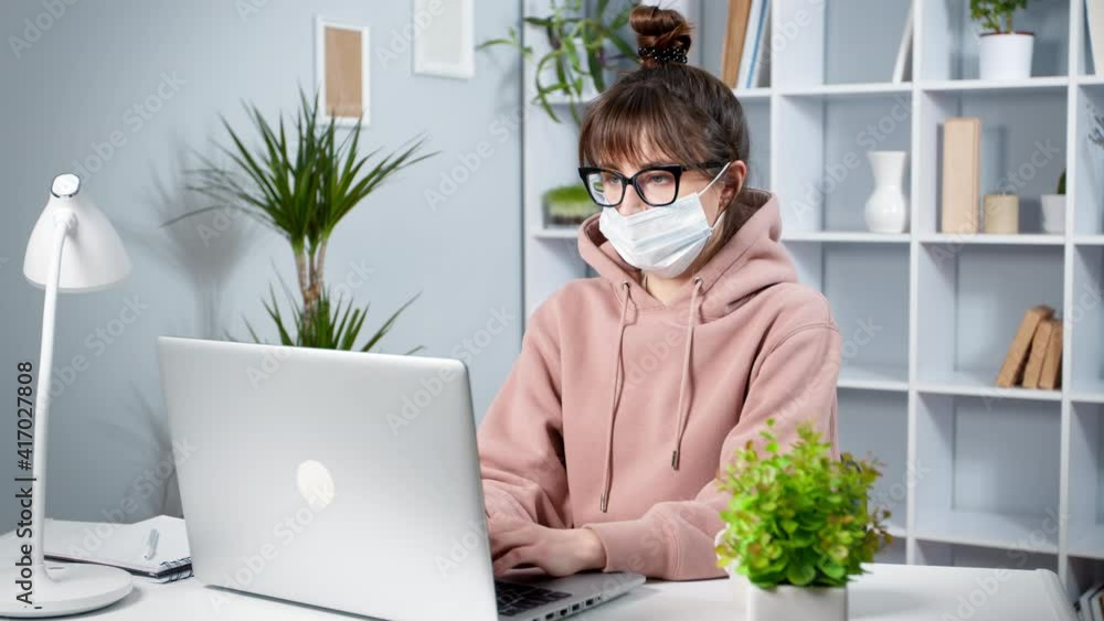 Young woman in protective mask works on laptop at home. Girl student is studying online. Young woman in protective mask works on laptop at home. Girl student is studying online.
