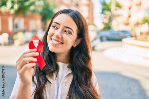 Фотография Young hispanic woman smiling happy holding red hiv ribbon walking at the city