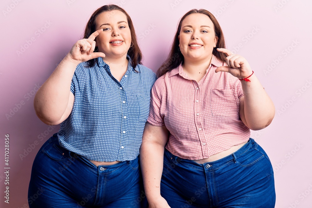 Young plus size twins wearing casual clothes smiling and confident gesturing with hand doing small size sign with fingers looking and the camera. measure concept. Young plus size twins wearing casual clothes smiling and confident gesturing with hand doing small size sign with fingers looking and the camera. measure concept.