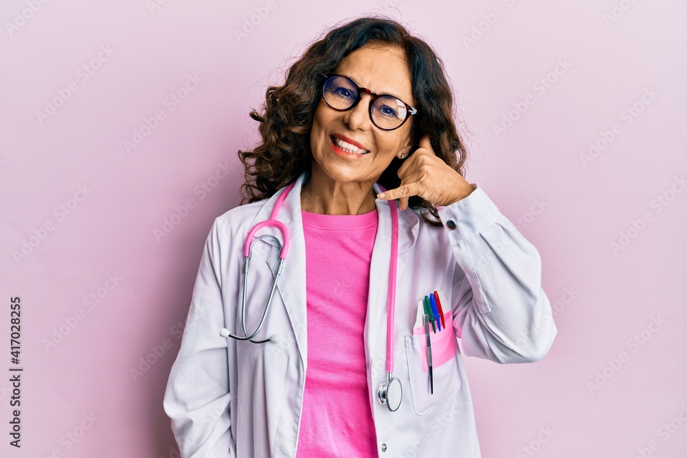Middle age hispanic woman wearing doctor uniform and glasses smiling doing phone gesture with hand and fingers like talking on the telephone. communicating concepts. Middle age hispanic woman wearing doctor uniform and glasses smiling doing phone gesture with hand and fingers like talking on the telephone. communicating concepts.