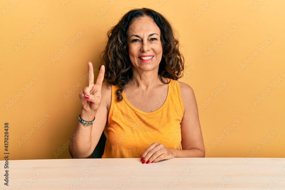 Beautiful middle age woman wearing casual clothes sitting on the table showing and pointing up with fingers number two while smiling confident and happy. Beautiful middle age woman wearing casual clothes sitting on the table showing and pointing up with fingers number two while smiling confident and happy.