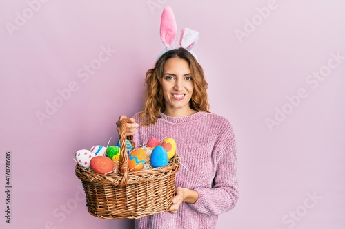 Canvas Print Young caucasian woman wearing cute easter bunny ears holding colored egg smiling with a happy and cool smile on face