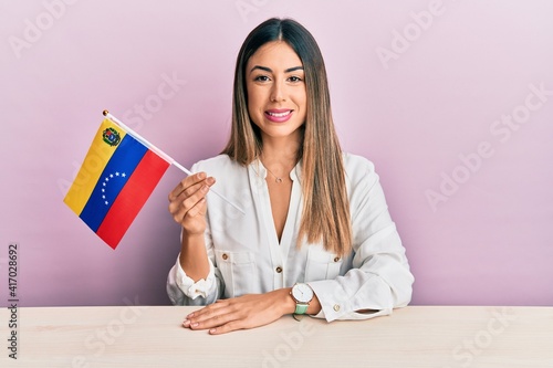 Young hispanic woman holding venezuelan flag sitting on the table looking positive and happy standing and smiling with a confident smile showing teeth photo