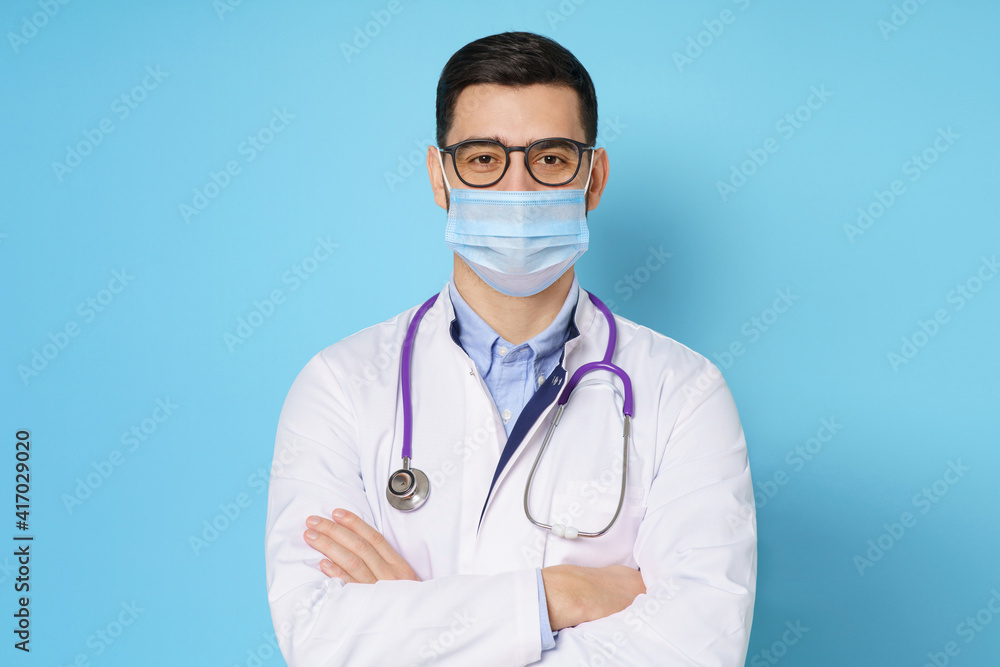 Young male doctor wearing medical mask, standing with crossed arms in white coat, isolated on blue background