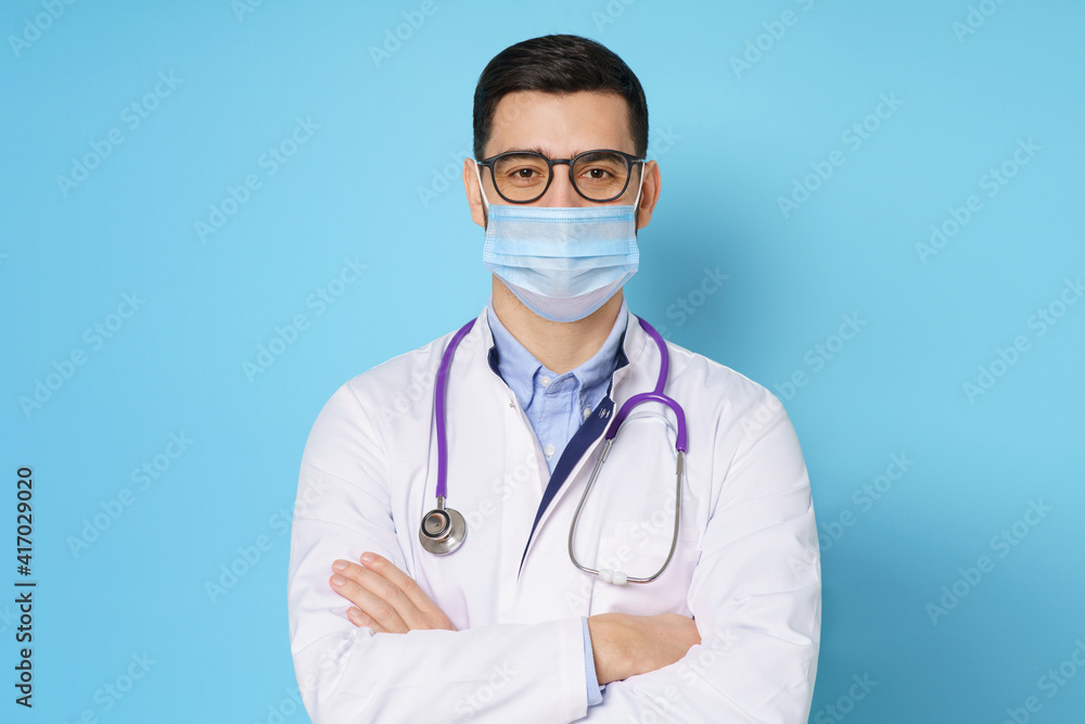 Young male doctor wearing medical mask, standing with crossed arms in white coat, isolated on blue background Young male doctor wearing medical mask, standing with crossed arms in white coat, isolated on blue background