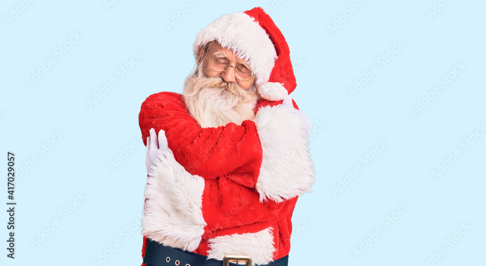 Old senior man with grey hair and long beard wearing traditional santa claus costume hugging oneself happy and positive, smiling confident. self love and self care
