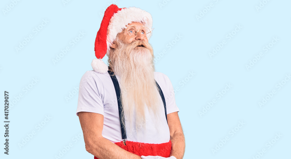 Obraz premium Old senior man with grey hair and long beard wearing santa claus costume with suspenders looking away to side with smile on face, natural expression. laughing confident.