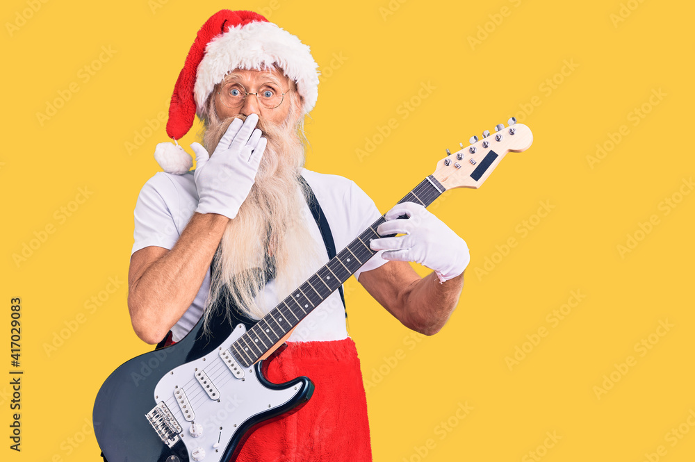 Old senior man with grey hair and long beard wearing santa claus costume playing electric guitar covering mouth with hand, shocked and afraid for mistake. surprised expression
