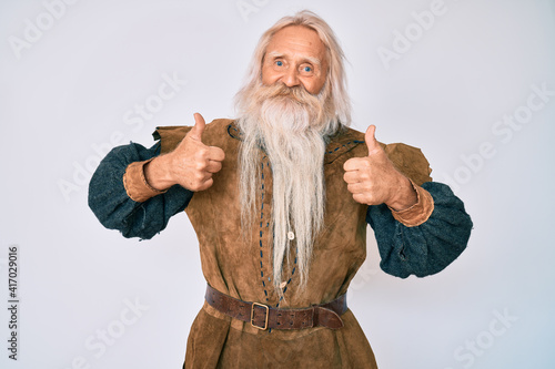 Old senior man with grey hair and long beard wearing viking traditional costume success sign doing positive gesture with hand, thumbs up smiling and happy. cheerful expression and winner gesture. photo