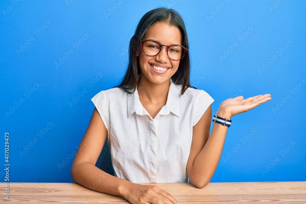 Beautiful hispanic woman wearing casual clothes sitting on the table smiling cheerful presenting and pointing with palm of hand looking at the camera. Beautiful hispanic woman wearing casual clothes sitting on the table smiling cheerful presenting and pointing with palm of hand looking at the camera.