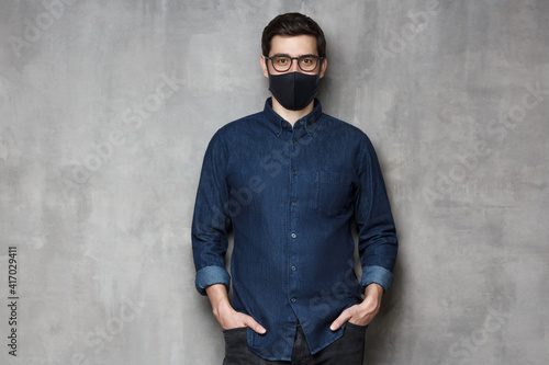 Young man wearing denim shirt, glasses and black mask, standing against gray textured wall