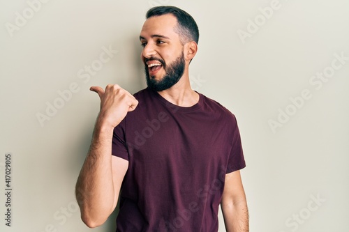 Young man with beard wearing casual t shirt pointing thumb up to the side smiling happy with open mouth