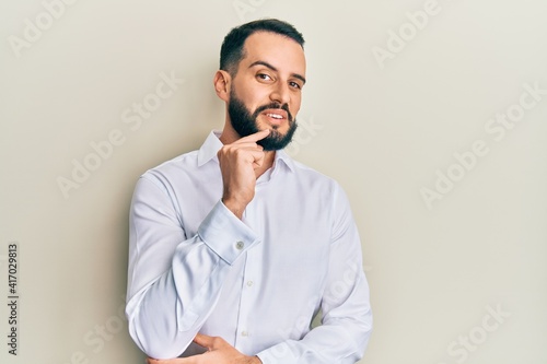Young man with beard wearing business white shirt smiling looking confident at the camera with crossed arms and hand on chin. thinking positive.
