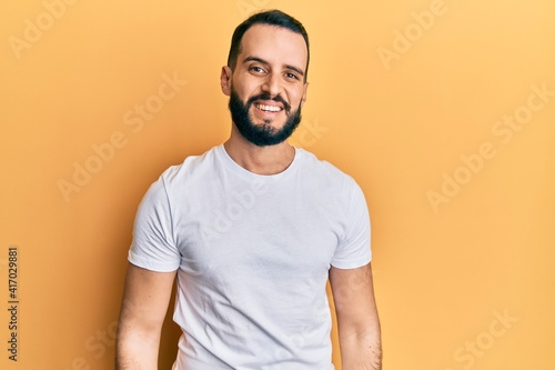 Young man with beard wearing casual white t shirt looking positive and happy standing and smiling with a confident smile showing teeth