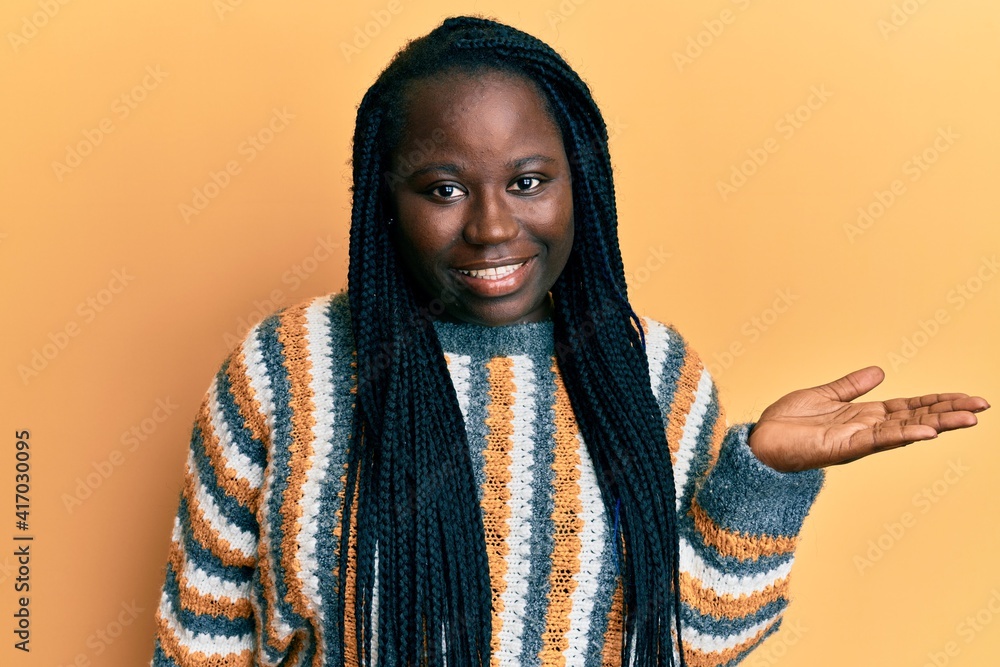 Young black woman with braids wearing casual winter sweater smiling cheerful presenting and pointing with palm of hand looking at the camera. Young black woman with braids wearing casual winter sweater smiling cheerful presenting and pointing with palm of hand looking at the camera.