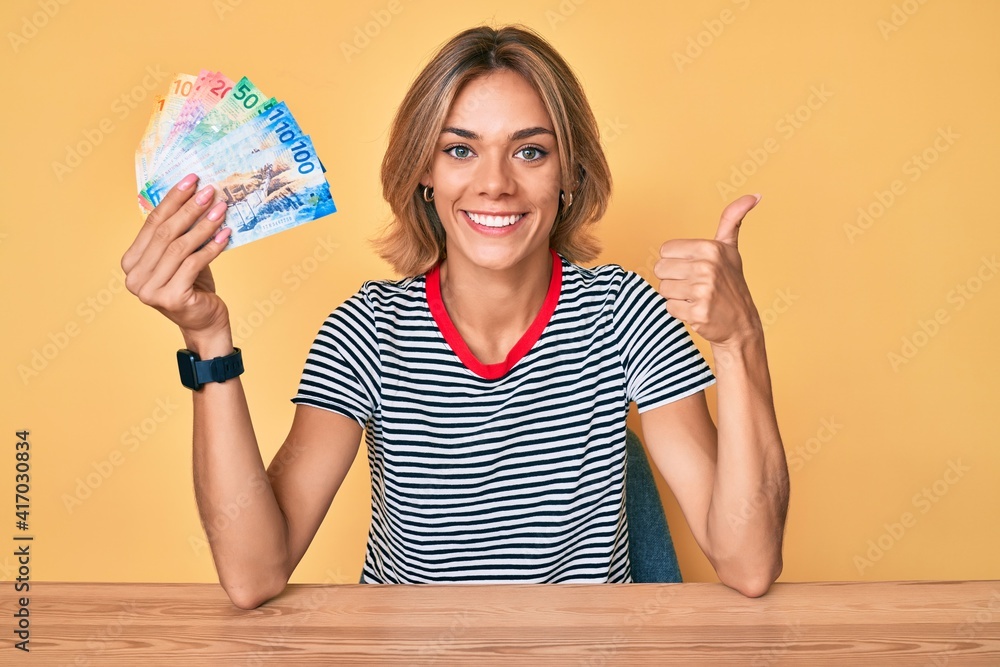Beautiful caucasian woman holding swiss franc banknotes smiling happy and positive, thumb up doing excellent and approval sign Beautiful caucasian woman holding swiss franc banknotes smiling happy and positive, thumb up doing excellent and approval sign