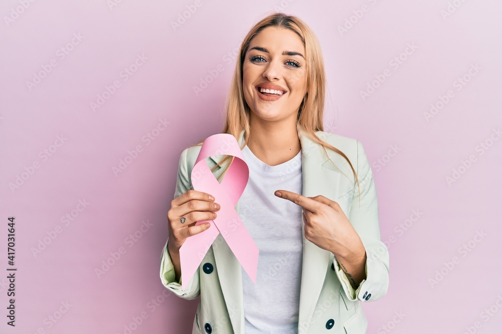 Young caucasian woman wearing business clothes holding pink cancer ribbon smiling happy pointing with hand and finger Young caucasian woman wearing business clothes holding pink cancer ribbon smiling happy pointing with hand and finger