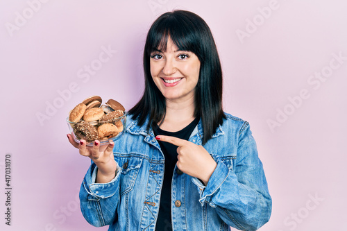 Young hispanic woman holding sweet pastries smiling happy pointing with hand and finger