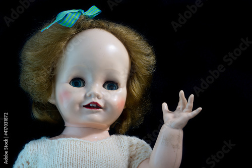 Konstfotografi Old vintage antique hard plastic doll with blond hair and staring eyes and waving an arm against a black background
