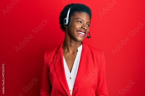 Fotografiet Young african american girl wearing call center agent headset looking away to side with smile on face, natural expression
