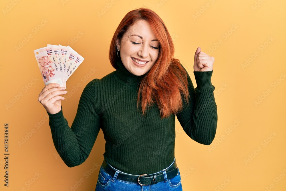 Beautiful redhead woman holding 10 singapore dollars banknotes screaming proud, celebrating victory and success very excited with raised arm Beautiful redhead woman holding 10 singapore dollars banknotes screaming proud, celebrating victory and success very excited with raised arm