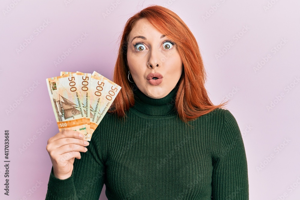 Beautiful redhead woman holding 500 norwegian krone banknotes scared and amazed with open mouth for surprise, disbelief face Beautiful redhead woman holding 500 norwegian krone banknotes scared and amazed with open mouth for surprise, disbelief face
