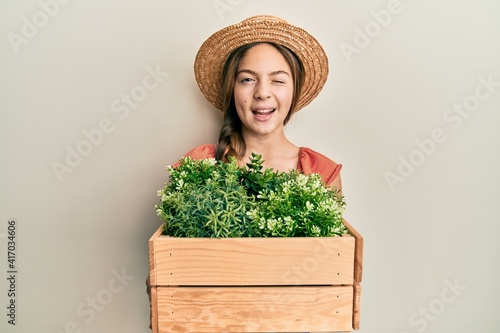 Valokuvatapetti Beautiful brunette little girl wearing gardener hat holding wooden plant pot winking looking at the camera with sexy expression, cheerful and happy face